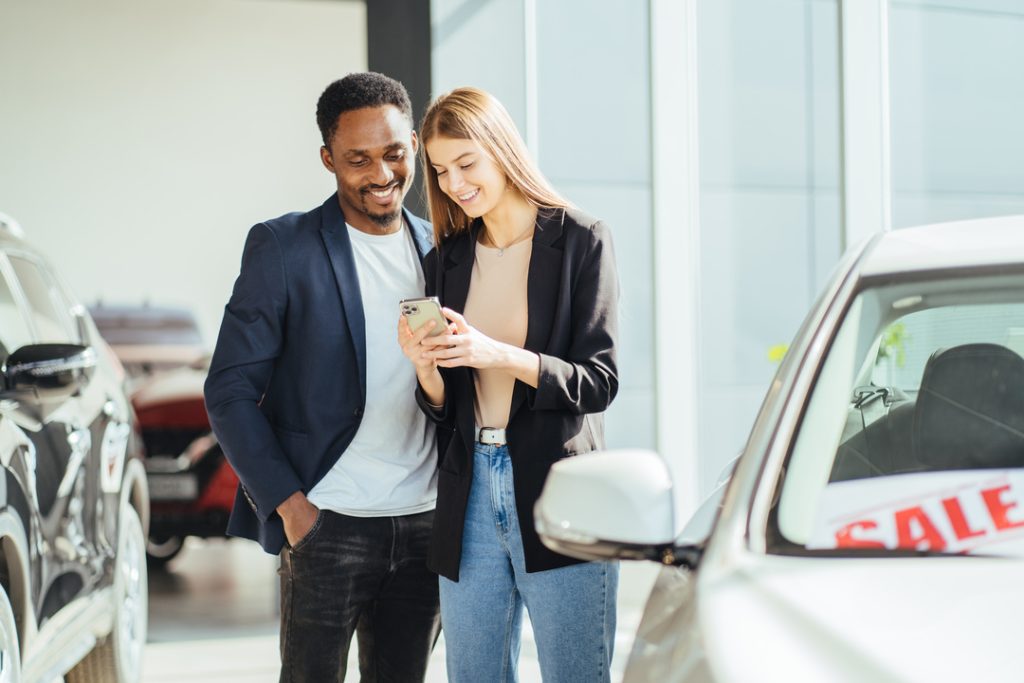 Multiracial couple using mobile while choosing car at salon - MotorPress Multiracial couple using mobile while choosing car at salon