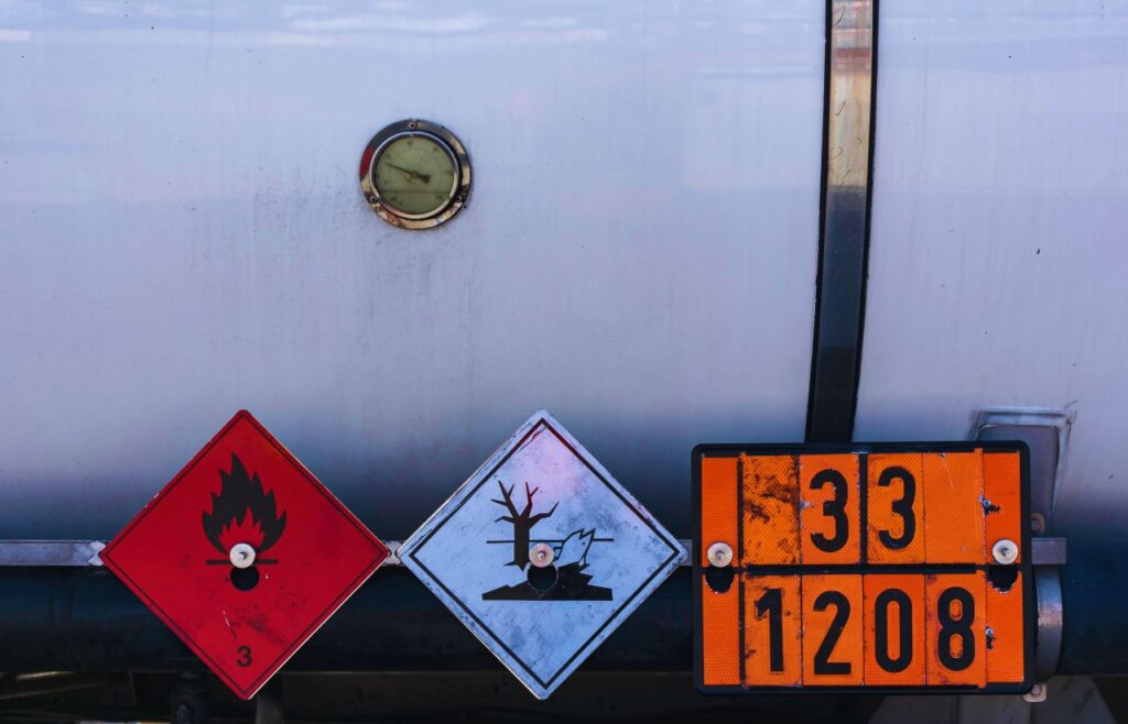 Rear of a tanker with three hazard placards: red diamond for flammable liquid (class 3), white diamond for environmental hazard (tree and dead fish), and an orange panel showing numbers 33 over 1208; gauge and tanker surface visible in background.
