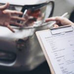Person holds a clipboard with a Personal Accident Claim Form beside a damaged car, ready to file a claim.