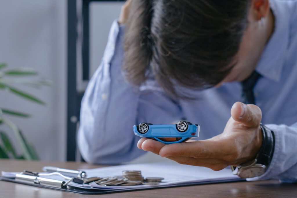 Businessperson in a blue shirt holds a small blue toy car over a desk with coins and financial documents nearby.