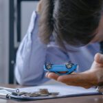 Businessperson in a blue shirt holds a small blue toy car over a desk with coins and financial documents nearby.
