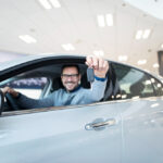 Man in a dealership showroom sits in a silver car, proudly holding up the car key.
