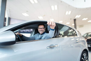 Man in a dealership showroom sits in a silver car, proudly holding up the car key.
