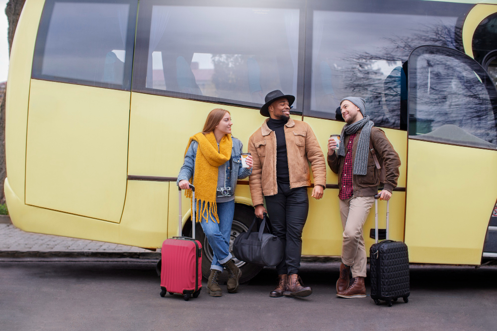 Three friends stand by a yellow bus with rolling suitcases, chatting and sipping drinks.