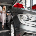 Couple walking beside a shiny new car in a showroom with a large red bow on the hood.