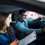 Man wearing glasses drives a car and smiles as a woman passenger holds a clipboard nearby.