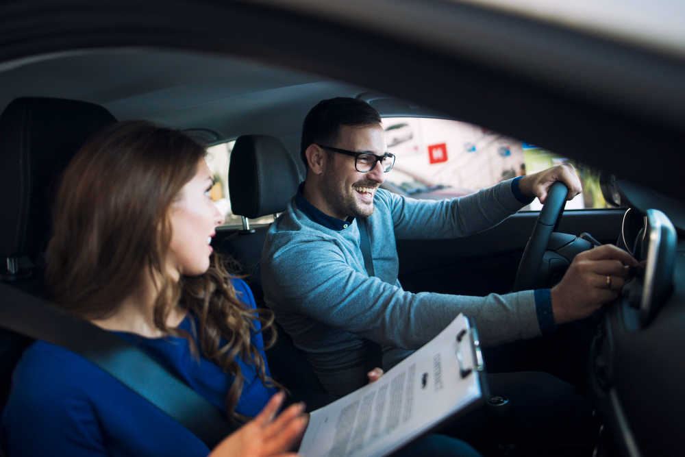Man wearing glasses drives a car and smiles as a woman passenger holds a clipboard nearby.