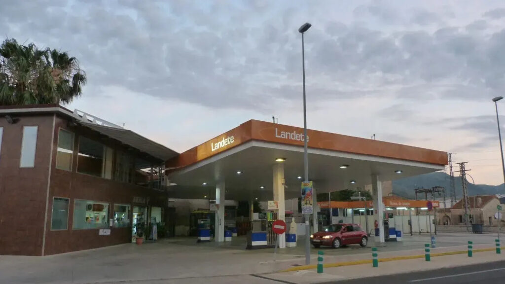 Gas station with an orange canopy labeled 'Landete' and a brick building nearby, under a cloudy sky.