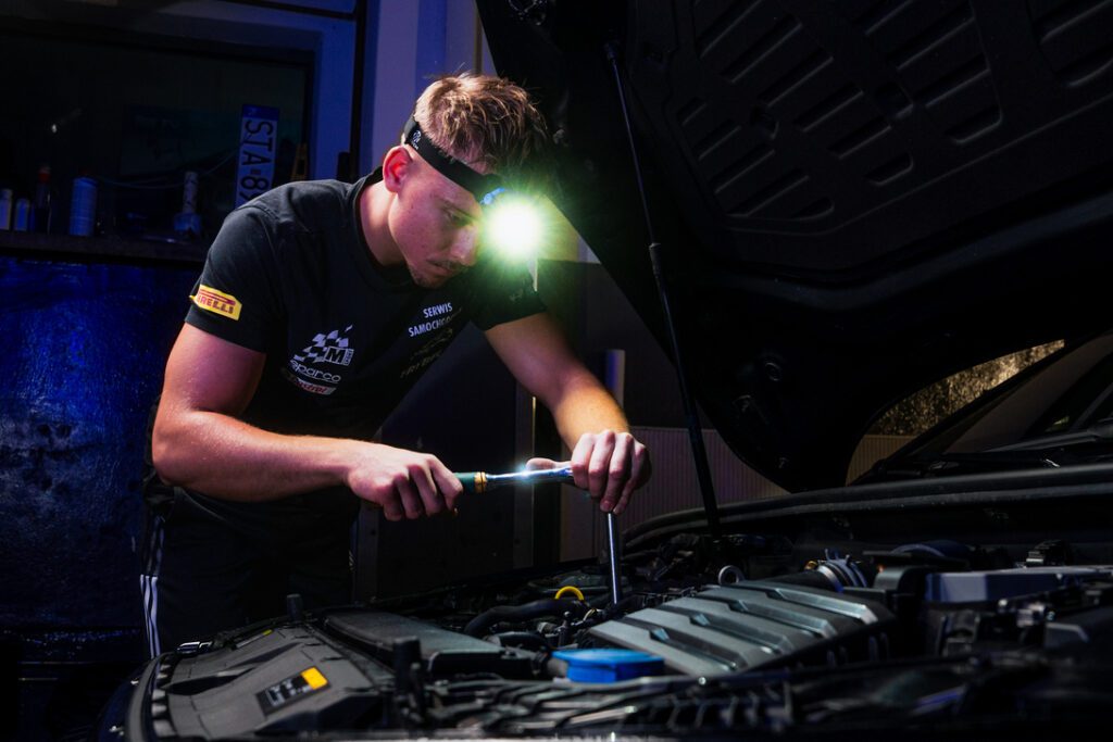 Mechanic wearing a headlamp and using a screwdriver to inspect a car engine under the hood in a dim workshop.