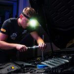 Mechanic wearing a headlamp and using a screwdriver to inspect a car engine under the hood in a dim workshop.