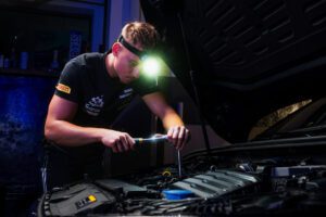 Mechanic wearing a headlamp and using a screwdriver to inspect a car engine under the hood in a dim workshop.