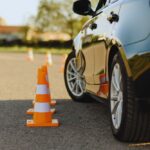 Car and a line of orange traffic cones set up on an asphalt parking lot, suggesting a driving course or drill.