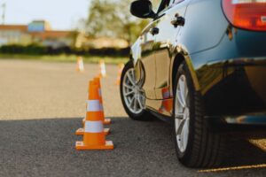 Car and a line of orange traffic cones set up on an asphalt parking lot, suggesting a driving course or drill.