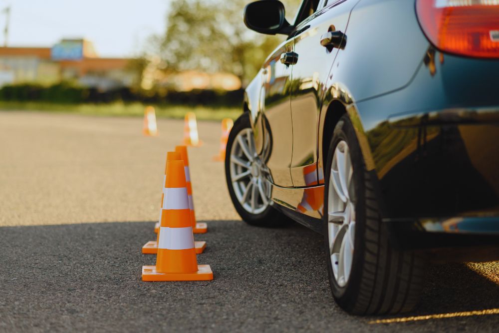 777944 - Autoescuela Isabel Segunda - MotorPress Car and a line of orange traffic cones set up on an asphalt parking lot, suggesting a driving course or drill.
