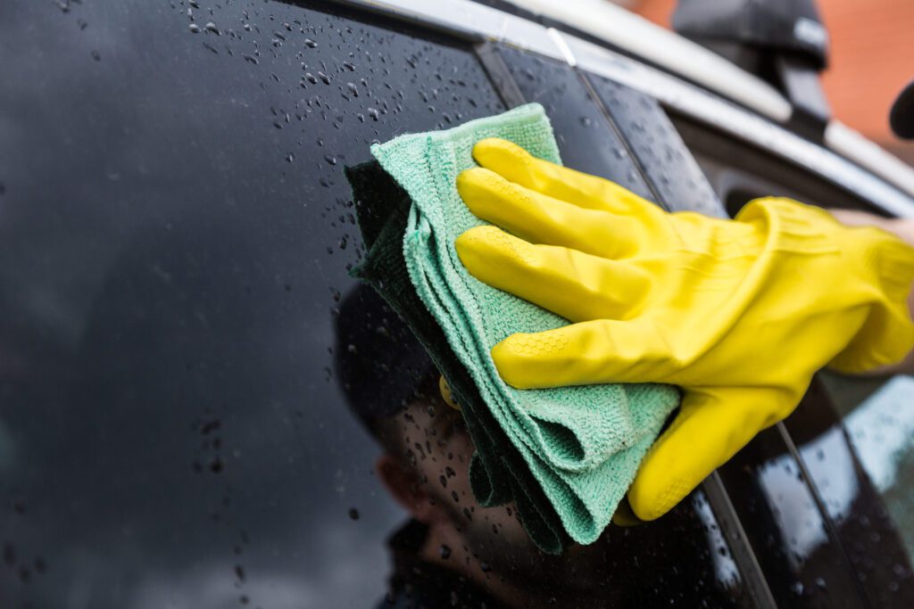 778201 Detailcar - MotorPress Person wearing yellow gloves wipes a wet car window with a green microfiber cloth, cleaning the surface.",
