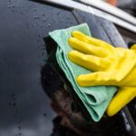 Person wearing yellow gloves wipes a wet car window with a green microfiber cloth, cleaning the surface.",