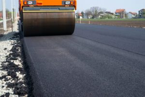Road roller compacting fresh asphalt on a newly paved rural road with gravel shoulder on the left and houses in the distance