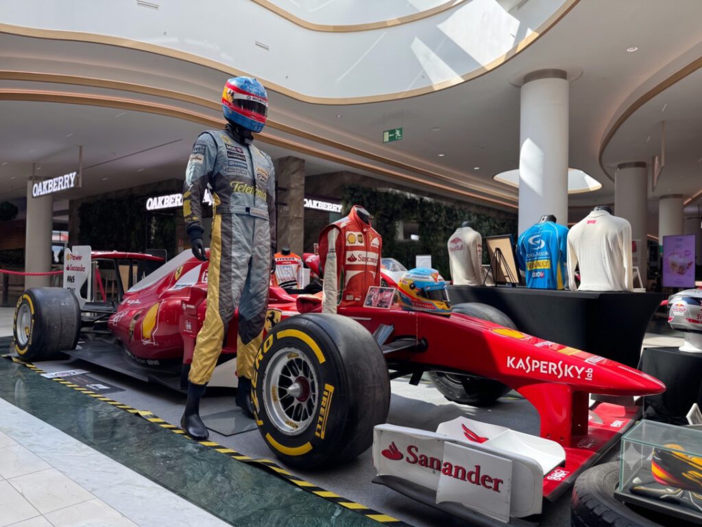 Red Formula 1 car on display in a shopping mall with a life-size driver mannequin in a racing suit beside it, branded with sponsors like Kaspersky and Sanander.