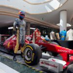 Red Formula 1 car on display in a shopping mall with a life-size driver mannequin in a racing suit beside it, branded with sponsors like Kaspersky and Sanander.