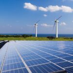Rows of solar panels stretching toward a grassy coastline with wind turbines and the ocean in the distance under a clear blue sky.
