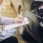 Man with a clipboard inspects the damaged rear bumper of a black car on a street.