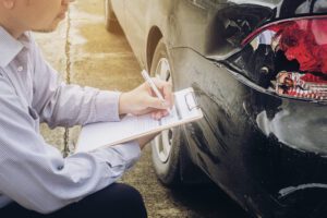 Man with a clipboard inspects the damaged rear bumper of a black car on a street.