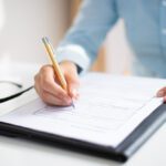Person in a blue shirt writes on a form with a pencil at a white desk, glasses resting nearby
