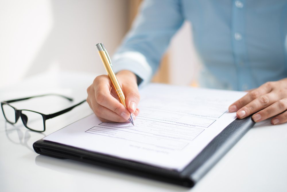 Person in a blue shirt writes on a form with a pencil at a white desk, glasses resting nearby