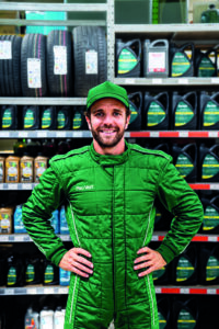 Man in a green racing suit stands with hands on hips in an auto parts store aisle.