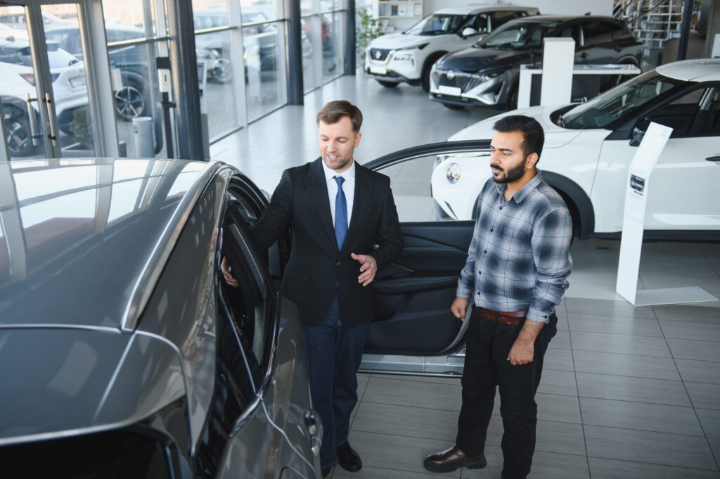 Sales consultant in a suit explains features to a customer beside an open car door in a car showroom.