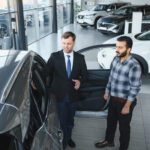 Sales consultant in a suit explains features to a customer beside an open car door in a car showroom.
