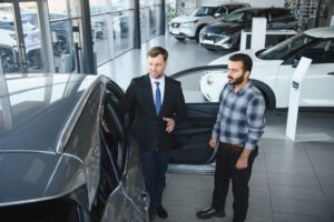Sales consultant in a suit explains features to a customer beside an open car door in a car showroom.